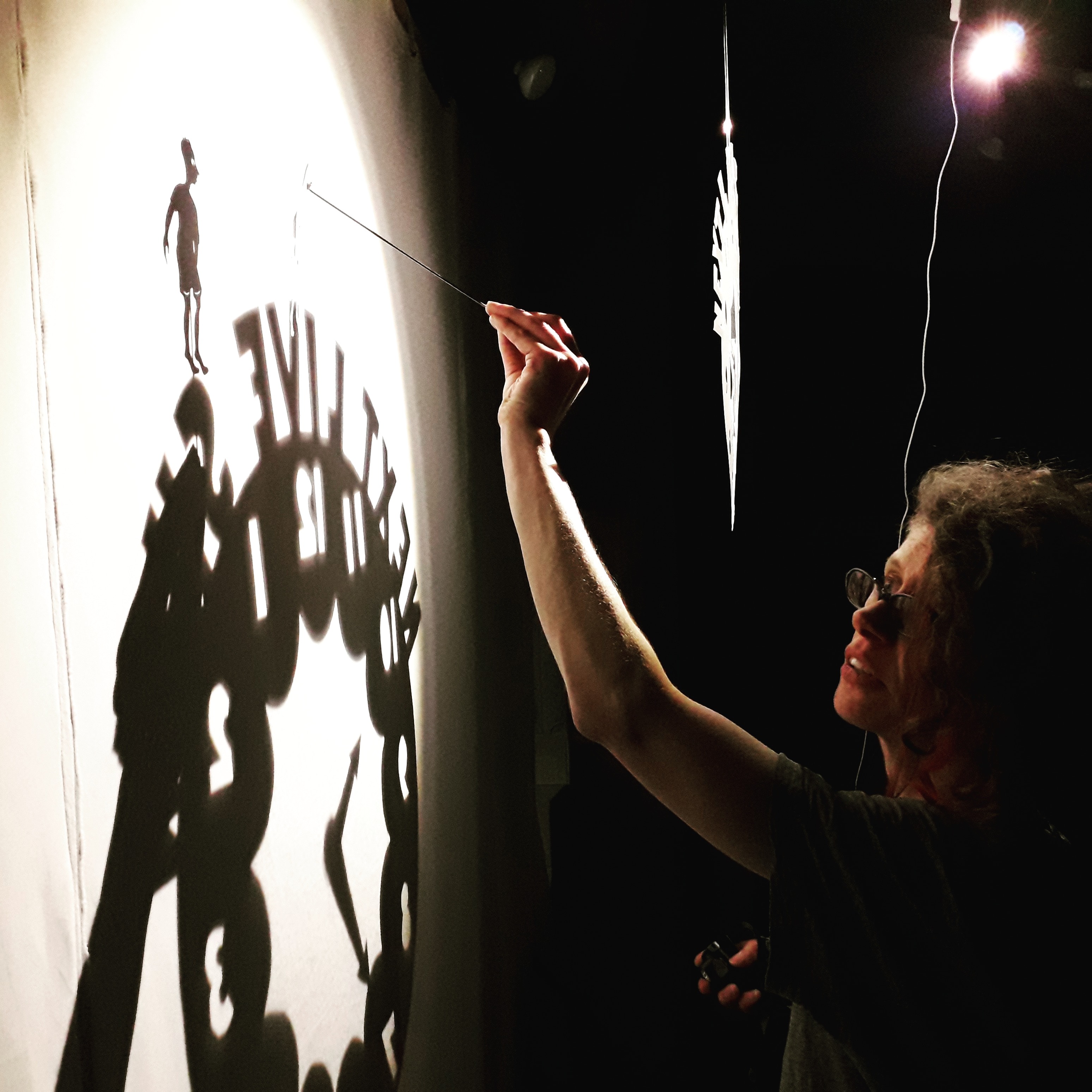 A darkened room with a single light source illuminates an image on a screen. A woman puppeteer is just visible in the darkness.